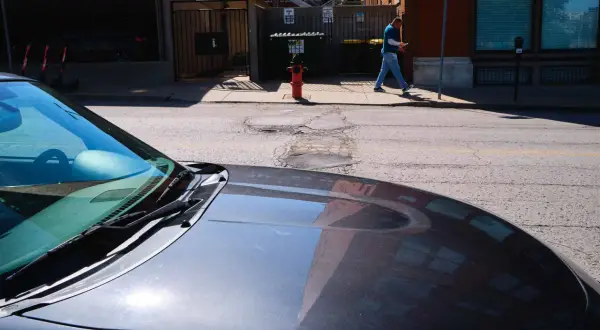 A man walks past a fire hydrant on a sunny day, his reflection visible in the hood of a car. 40mm street photography captures the urban scene.