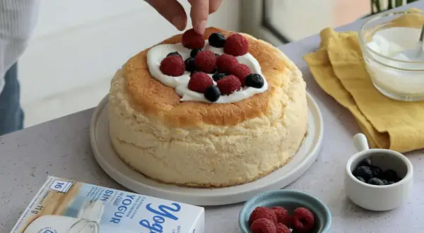 Tarta de queso esponjosa decorada con frutos rojos y nata. Se ve una caja de yogur griego al lado.