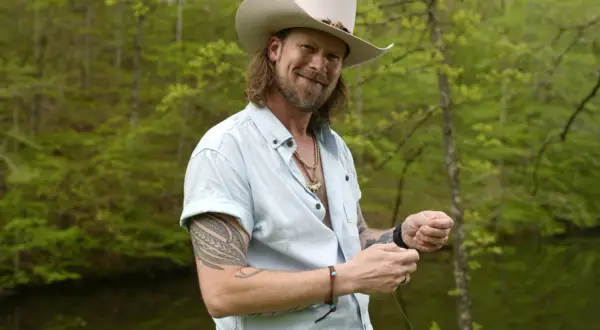 Brian Kelley, in a cowboy hat and light blue shirt, smiles while holding fishing line, perhaps inspired by his songs.