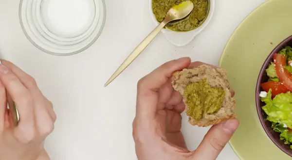 Main d'une personne tenant un morceau de pain avec du pesto, à côté d'une salade.