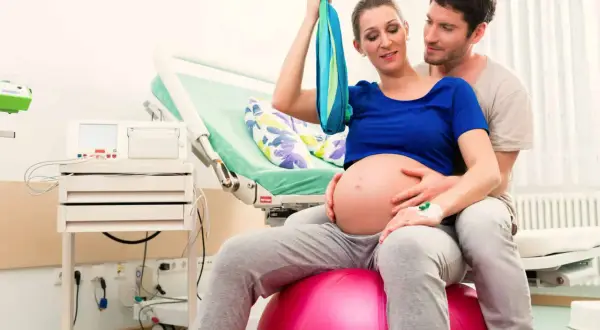Pareja en sala de partos. Ella sentada en balón, buscando la mejor postura para parir, él la abraza.