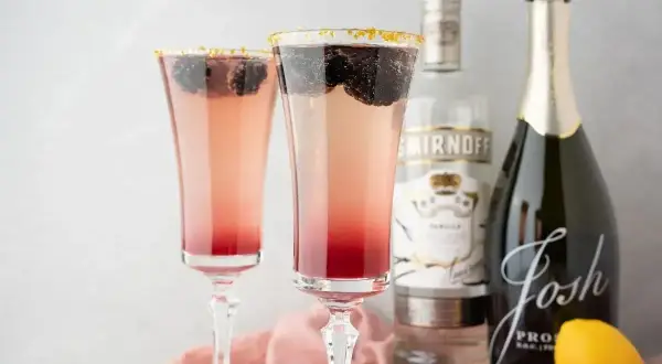 Dwa eleganckie drinki z prosecco, ozdobione jeżynami i złotym cukrem, obok butelek prosecco i wódki.