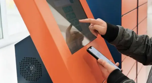Osoba korzysta z ekranu dotykowego automatu, by dowiedzieć się, jak wygenerować etykietę InPost. Trzyma też telefon.