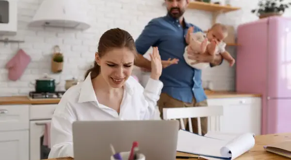 Zestresowana kobieta przy laptopie, mąż z dzieckiem w tle. Czy przerwa na karmienie piersią do jakiego wieku dziecka jest możliwa w takiej sytuacji?