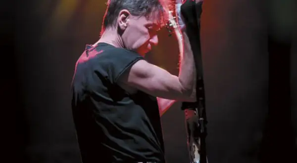 Muzyk w czerni, z czerwoną poświatą na włosach, przygotowuje się do wykonania swoich ksu utworów na gitarze.