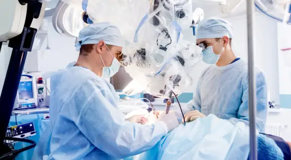 Chirurdzy w maseczkach i fartuchach operacyjnych pracują przy użyciu mikroskopu.
