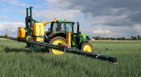 Zielony traktor z opryskiwaczem polowym, gotowy do pracy z adiuwantem, na tle pola i chmur.