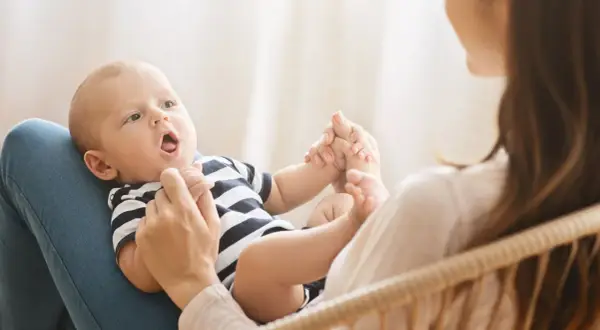 Ein Baby mit offenem Mund liegt auf dem Schoß seiner Mutter. Es hält ihre Hände. Bald sagt das Baby "Mama".