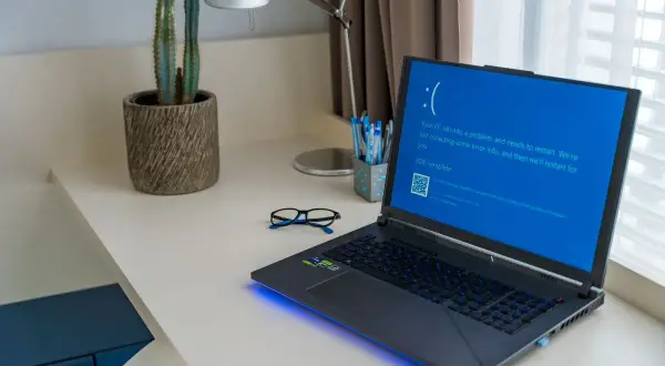 Laptop z niebieskim ekranem śmierci, wymagający twardy reset laptopa. Na biurku okulary i kaktus.