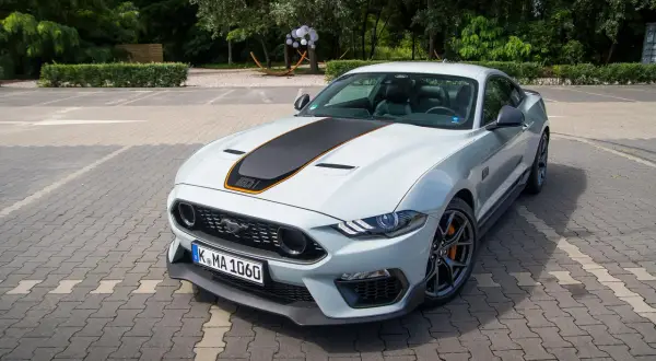 Biały Ford Mustang Mach 1 z czarnym pasem na masce i pomarańczowymi akcentami, zaparkowany na kostce brukowej.