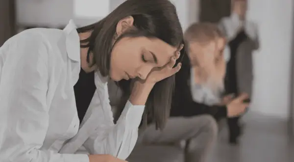 En ung kvinna med handen mot pannan, kanske i väntan på en **social fobi diagnos**. I bakgrunden syns andra personer.