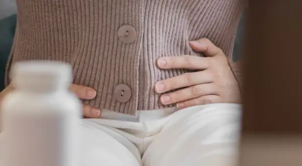 Kobieta trzyma się za brzuch, szukając ulgi na wzdęcia. Obok stoi biała butelka z lekami.