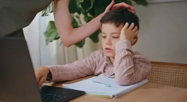 Chłopiec z trudem uczy się efektywnie przy laptopie, a rodzic wspiera go, kładąc dłoń na głowie.