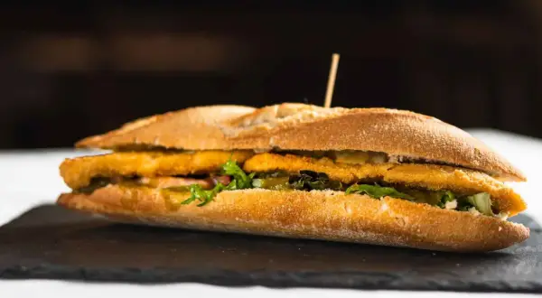 Un bocadillo caliente con rebozado crujiente, lechuga fresca y pepinillos, servido en pan rústico.