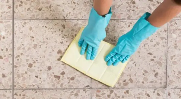 Manos con guantes azules limpiando suelos de terrazo con un paño amarillo.