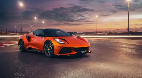 Pomarańczowy Lotus Emira 420 Sport na torze wyścigowym o zachodzie słońca, z rozmazanymi światłami w tle.