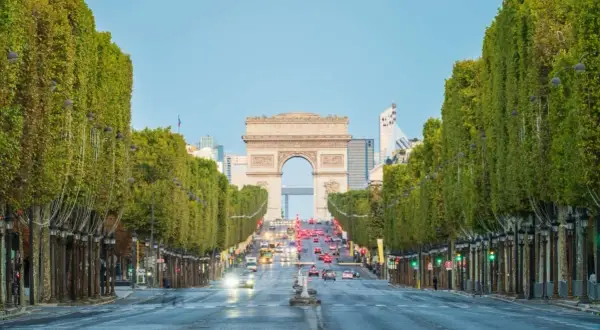 Aleja **Champs-Élysées** w Paryżu z Łukiem Triumfalnym w oddali. Drzewa tworzą zielony tunel, a ruch samochodowy płynie w stronę monumentu.