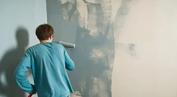 Un uomo con una maglietta blu sta dipingendo un muro con un rullo. La pittura si stacca dal muro in alcune zone, creando un effetto astratto.