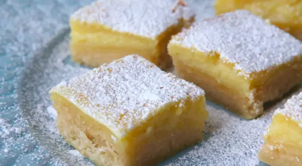 Färska bitar av citronkaka i långpanna, pudrade med florsocker, ligger på en blå tallrik.