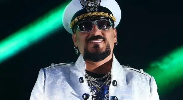 Gigi D'Agostino w białej marynarce i czapce, uśmiechnięty. Mimo choroby, jego muzyka wciąż porusza serca fanów.