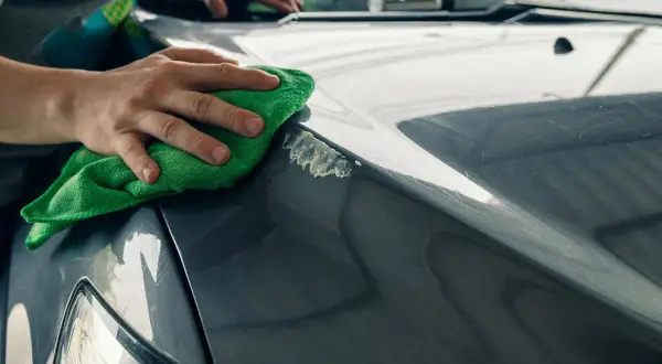 Manos puliendo el capó de un coche gris con un paño verde, mostrando cómo quitar arañazos del coche.