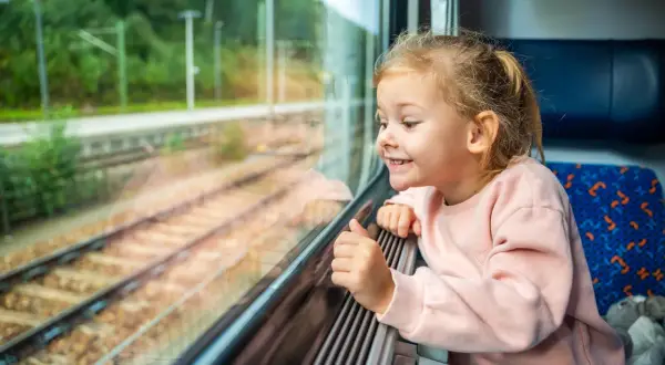 Uśmiechnięta dziewczynka wpatruje się przez okno pociągu. Zastanawia się, od ilu lat można jeździć samemu pociągiem, obserwując mijane krajobrazy.