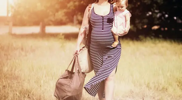 Schwangere Frau mit Baby im Arm und Taschen, die sich durch hohes Gras kämpft. Schwer heben schwangerschaft.