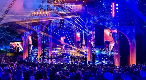 Koncertowa scena z tłumem widzów, oświetlona niebieskimi i różowymi światłami, z ekranami wyświetlającymi obrazy i wieżę Eiffla.