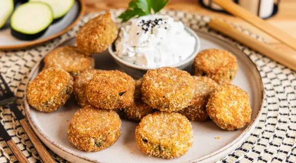 Knusprig panierte Zucchini-Taler auf einem Teller, serviert mit einem Dip und garniert mit Petersilie. Ein leckerer Snack!