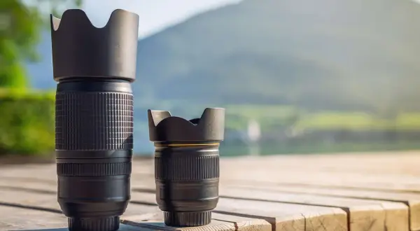 Two camera lenses, a tall telephoto and a shorter normal lens, sit on a wooden deck with a blurred mountain and lake background.
