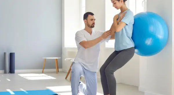Fizjoterapeuta pomaga kobiecie w ćwiczeniu aktywującym mięśnie głębokie przy użyciu piłki gimnastycznej.