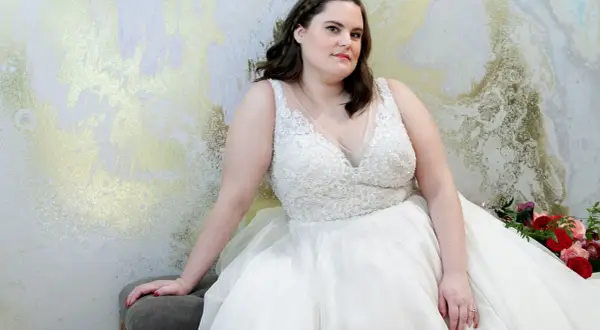 Kobieta w białej, zdobionej sukni ślubnej plus size siedzi na tle złoto-białej ściany.