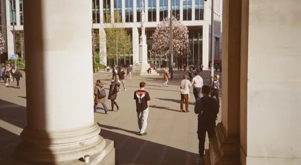 A 35mm street photography scene captures people walking in a plaza with a modern building and blooming trees.