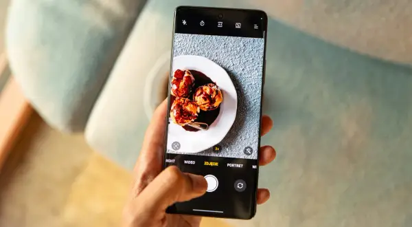 Dłoń trzyma smartfon z otwartą aplikacją do robienia zdjęć. Na ekranie widoczny jest talerz z trzema ptysiami polanymi czekoladą.