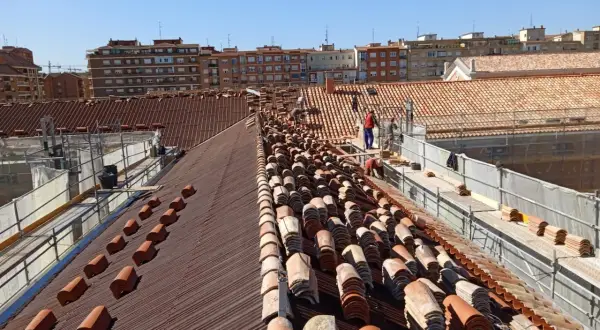 Trabajadores renuevan las cubiertas de tejas de un edificio, con pilas de tejas nuevas y viejas a la vista.