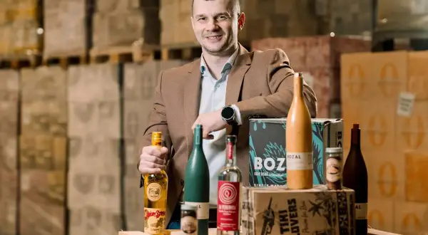 Mężczyzna w brązowej marynarce prezentuje butelki z alkoholem, w tym te z etykietą "Spice Bros". W tle magazyn pełen towarów, sugerujący dystrybucję california alkohol.
