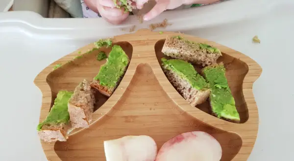 Babyhände greifen nach Avocado-Brotstücken auf einem Holzbrett. Ein 10 Monate altes Baby erkundet sein Essen.