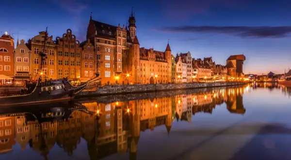 Gdańsk nocą, z zabytkowymi kamienicami i żaglowcem odbijającymi się w spokojnej wodzie Motławy.