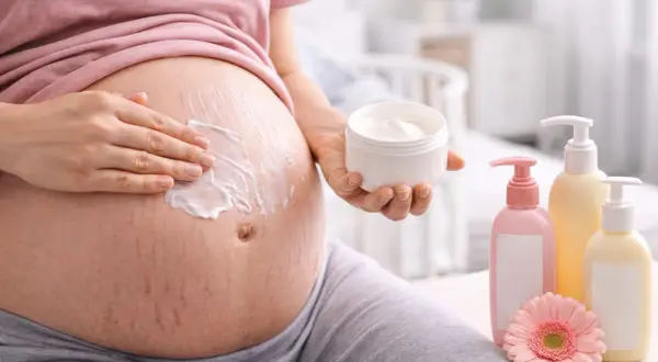 Ciężarna kobieta smaruje brzuch kremem. W pobliżu stoją butelki z kosmetykami i kwiat. Bezpieczne dla skóry w ciąży, np. bez kwasu glikolowego.