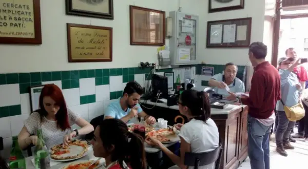 Wnętrze najstarszej pizzerii w Neapolu. Ludzie jedzą pizzę, a za ladą obsługuje kelner.