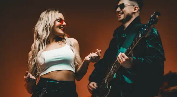 Para z uśmiechem, ona w czerwonych okularach, on gra na gitarze. Ich dłonie się stykają, jakby tworzyły tekst.