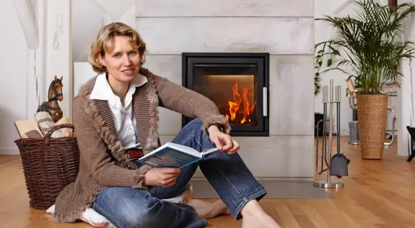 Frau liest Buch vor Kaminofen in Mietwohnung. Holzkorb daneben.