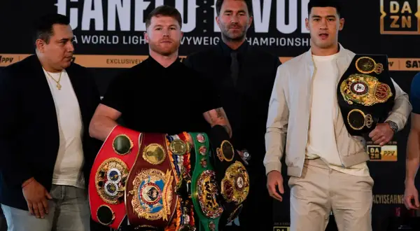 Canelo Alvarez, surrounded by championship belts, shows how good he is. He stands with Dmitry Bivol and Eddie Hearn.