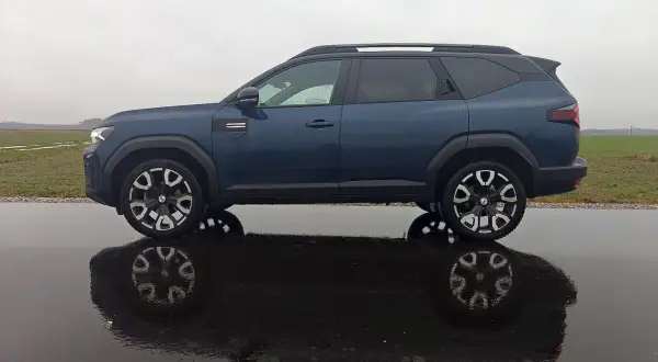 Dacia Bigster LPG w ciemnoniebieskim kolorze, zaparkowana na mokrej drodze, z odbiciem w kałuży.