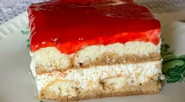 Kawałek ciasta bez pieczenia z warstwami biszkoptu, kremu i galaretki truskawkowej na talerzyku.