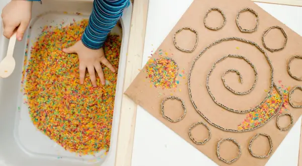 Main colorée dans un bac, une main d'enfant explore. À côté, un carton en spirale avec des ronds découpés, parsemé de riz coloré. Une activité sensorielle adulte pour stimuler la créativité.