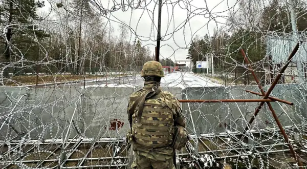 Żołnierz w mundurze obserwuje teren przez zasieki z drutu kolczastego. Widoczna jest granica polsko-białoruska, z budynkami i znakami po drugiej stronie.
