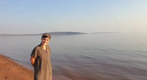 Mujer sonriente en la orilla de arena rojiza del lago Kariba, con el agua tranquila y el cielo despejado al fondo.