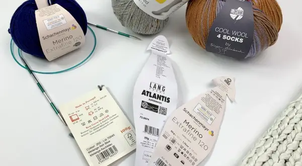 Wollknäuel zeigen den Unterschied zwischen Schurwolle und Wolle. Ein blauer Merino-Strang und ein Tweed-Garn liegen neben Stricknadeln.