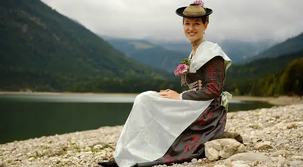 Frau in traditioneller Isarwinkler Tracht sitzt am Ufer eines Sees vor einer Bergkulisse.